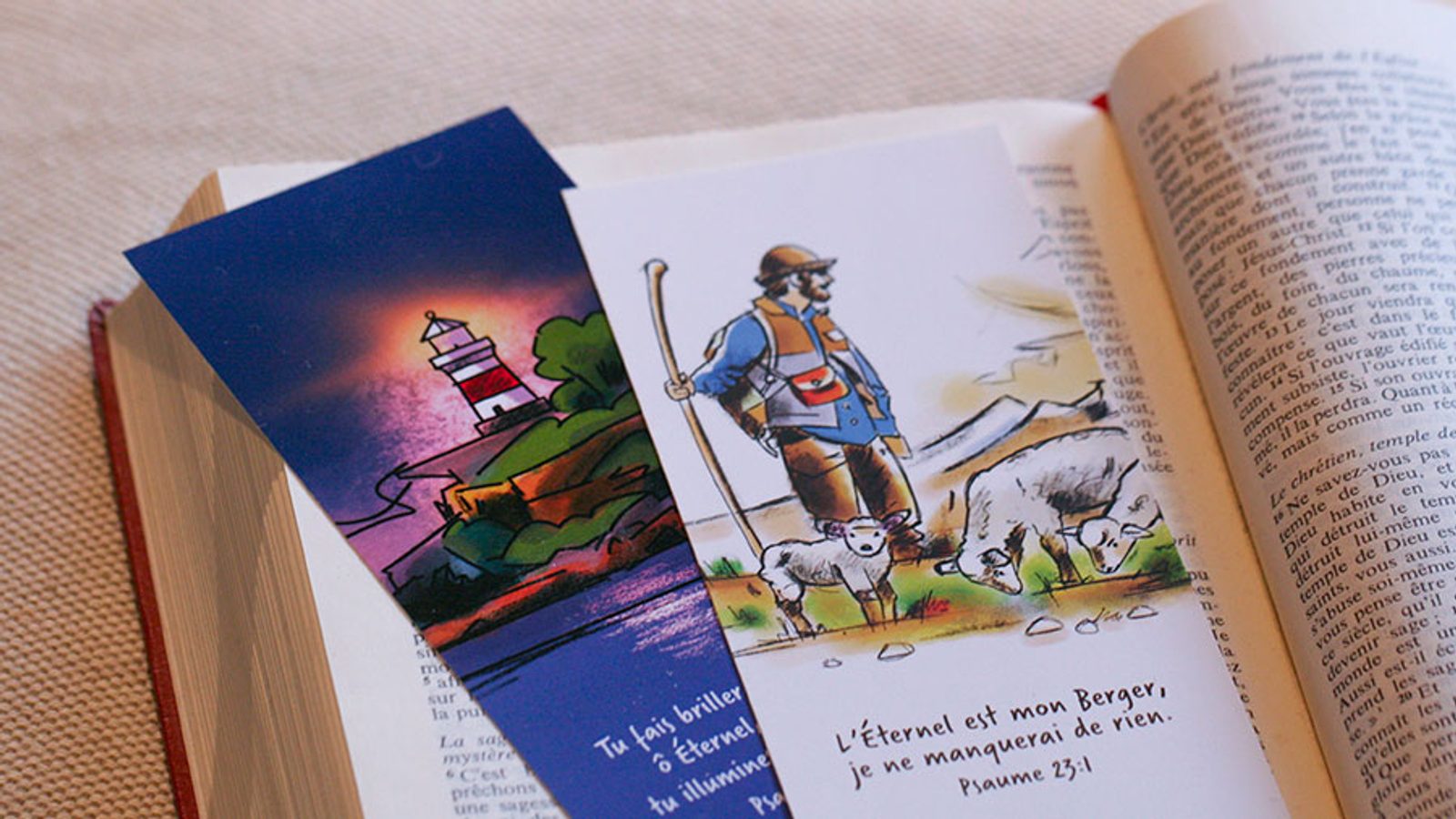 Photo d’une Bible ouverte accompagnée de deux signets illustrés représentant un phare et le Bon Berger, symboles de guidance, de lumière et de foi chrétienne.